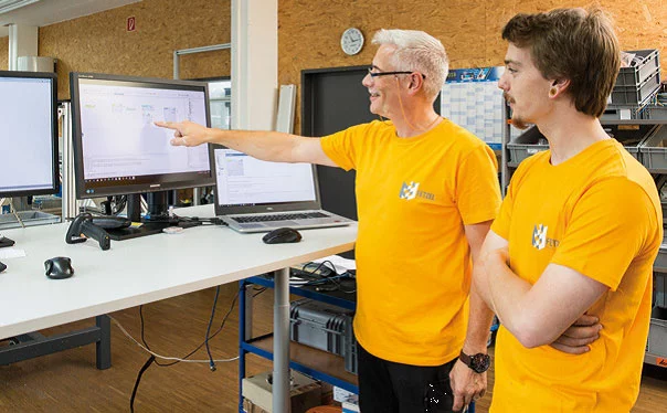 Zeigt zwei Mitarbeiter aus der Elektroabteilung. Der eine zeigt dem anderen etwas auf dem Bildschirm.